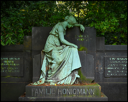 Bronzegrüne Skulptur einer sitzenden, lesenden Frau auf dem Grabmal der Familie Honigmann. Dunkle Steintafeln mit Inschriften im Hintergrund, Herbstlaub am Boden.