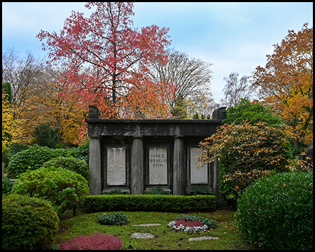 Modernes, dunkles Mausoleum im Bauhausstil mit drei rechteckigen Nischen. Herbstlich gefärbte Bäume in Rot und Orange dahinter. Gepflegte Buchsbaumhecken und violettes sowie rotes Heidekraut davor.