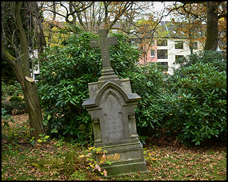 Neugotischer, verwitterter Grabstein mit spitzbogiger Verzierung und Kreuz-Motiv. Umgeben von Rhododendren und Herbstlaub. Im Hintergrund moderne Gebäude.