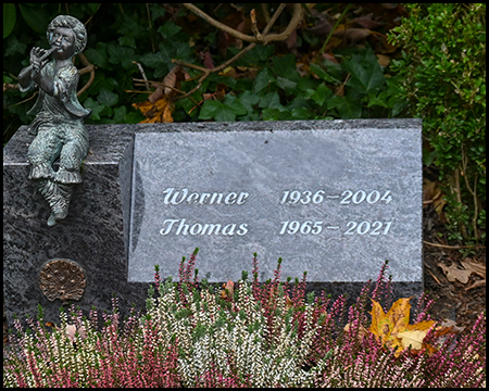 Moderne Grabplatte aus grauem Stein mit den Namen "Werner 1936-2004" und "Thomas 1965-2021". Links eine kleine Bronzefigur eines sitzenden Kindes. Davor buntes Heidekraut und ein gelbes Ahornblatt.