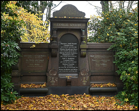 Großes, dunkles historisches Familiengrabmal mit der Aufschrift "Familie Knaudt", geschmückt mit Bogennische und seitlichen Tafeln. Herbstlaub liegt auf den Seitenflügeln. Gelb leuchtende Bäume im Hintergrund.