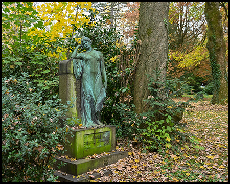 Bronzegrüne Skulptur einer verschleierten Trauerfigur auf moosbedecktem Steinsockel. Die Figur steht zwischen Baumstämmen, umgeben von Efeu und Herbstlaub auf dem Boden.