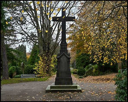 Hohes dunkles Steinkreuz auf mehrstufigem Sockel an einer Friedhofskreuzung. Herbstlich gefärbte Bäume in Gelb und Grün im Hintergrund, Laub liegt auf dem asphaltierten Weg.