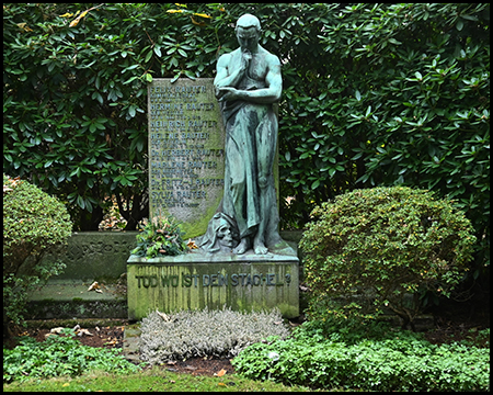 Bronzegrüne nachdenkliche Skulptur mit zu einem Schädel hinabgeneigtem Kopf auf Steinsockel. Dahinter eine Gedenktafel. Angebrachter Satz "Tod, wo ist dein Stachel?" Umgeben von akkurat geschnittenen Büschen und dichtem Grün.