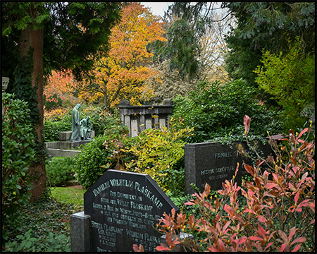 Friedhofsszene mit verschiedenen Grabsteinen im Vordergrund, darunter ein dunkler Stein. Im Hintergrund herbstlich gefärbte Bäume, Skulpturen und ein Mausoleum. Rotbraune Sträucher rechts im Bild.