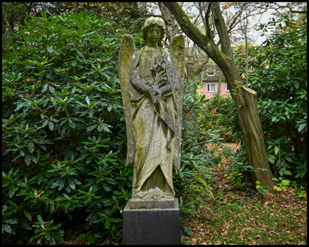 Verwitterte Steinskulptur einer Frauenfigur mit Blumen im Arm auf dunklem Sockel. Die moosbedeckte Figur steht zwischen Rhododendren, im Hintergrund ein rotes Wohnhaus und Herbstlaub am Boden.