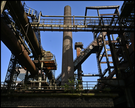 Gegen den blauen Himmel sichtbare Bauteile einer Industrieanlage mit Leitungen, Rohren und Schornstein