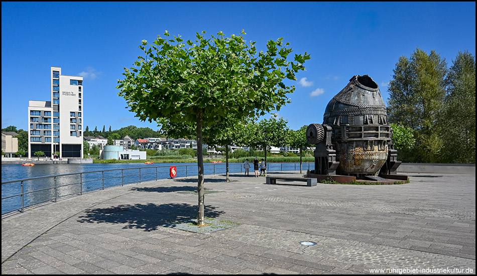 Moderne Uferpromenade mit grünem Baum, Wasserfläche und einem beeindruckenden industriellen Metallobjekt, das wie ein alter Konverter aussieht. Im Hintergrund ein modernes Hochhaus unter blauem Himmel.