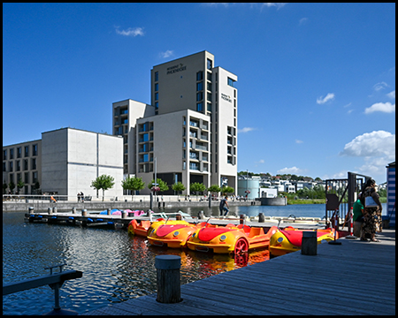 Bunte Tretboote in Form von Autos liegen am Anlegesteg eines Sees. Im Hintergrund steht ein modernes weißes Hochhaus, das sich im ruhigen Wasser spiegelt.