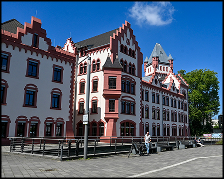 Historisches Gebäude mit roter Fassade und dekorativen Giebeln im Stil der Neugotik. Das imposante Bauwerk liegt direkt am Ufer und verfügt über eine charakteristische Architektur mit hellen Akzenten.
