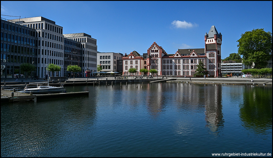 Blick über einen ruhigen See auf ein historisches Gebäude mit rot-weißer Fassade und Turmelementen. Im linken Bildbereich moderne Bürogebäude, im Vordergrund ein paar Boote am Steg.