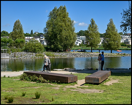 Erholungsbereich am Seeufer mit Sitzbänken aus
Stahl. Im Hintergrund eine Wasserfläche mit kleiner Halbinsel und dahinter Wohnbebauung auf einer Anhöhe unter blauem Himmel.