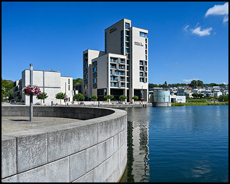 Modernes Hochhaus mit auffälligem Treppenturm spiegelt sich im Wasser. Im Vordergrund eine geschwungene Uferbefestigung aus Beton