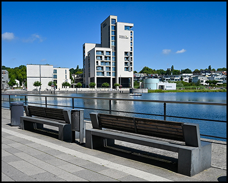 Blick über eine Sitzecke mit modernen Bänken auf ein markantes weißes Hochhaus am Ufer eines Sees. Die minimalistische Architektur kontrastiert mit der Naturumgebung.
