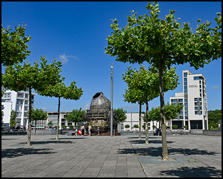 Gepflasterter Platz mit gleichmäßig gepflanzten jungen Bäumen und einer industriellen Metallskulptur in der Mitte. Im Hintergrund ein modernes Hochhaus und Wasserfläche