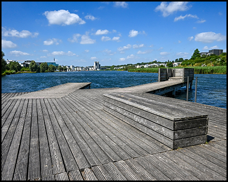 Holzsteg, der ins Wasser führt, mit einer Holzbank im Vordergrund. Blick über den See auf die Stadtsilhouette unter blauem Himmel mit weißen Wolken