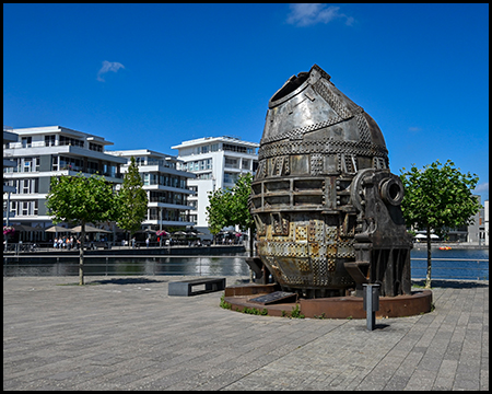 Nahaufnahme eines beeindruckenden historischen Metallkonverters, der als Industriedenkmal aufgestellt wurde. Im Hintergrund moderne Wohngebäude am Wasser.