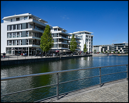 Moderne weiße Wohngebäude am Ufer eines Sees mit gepflegter Promenade. Die Architektur ist geprägt von großen Fensterfronten und Balkonen