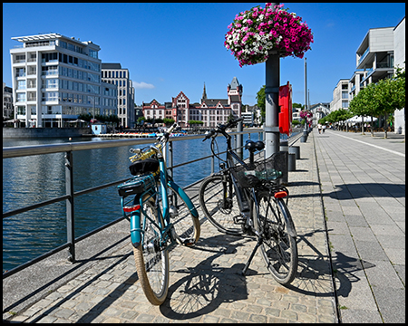 Fahrräder an einem Geländer entlang der Uferpromenade mit bunten Blumenampeln. Im Hintergrund ein historisches Gebäude mit charakteristischer rot-weißer Fassade am anderen Ufer des Gewässers