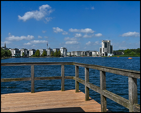 Holzsteg mit Geländer, der einen Blick über das Wasser auf die moderne Stadtsilhouette bietet. Wohngebäude und ein markanter Turm zeichnen sich am anderen Ufer ab.