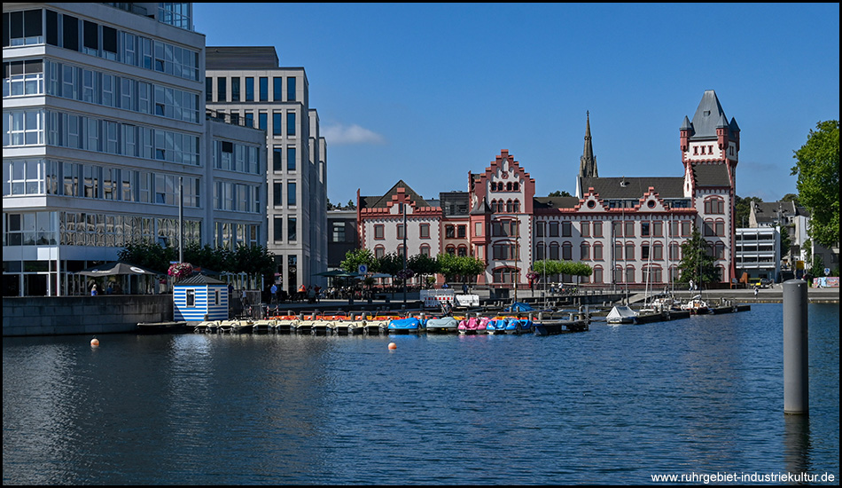 Blick über das Wasser auf eine Mischung aus modernen und historischen Gebäuden. Im Vordergrund ein kleiner Bootssteg mit bunten Tretbooten, im Hintergrund ein beeindruckendes Gebäude mit rot-weißer Fassade und Turm.