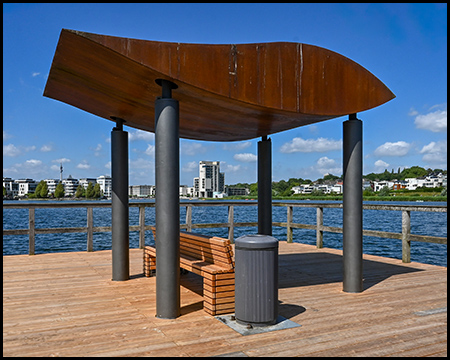 pavillon mit geschwungenem Dach auf Metallsäulen an einem Anlegesteg. Darunter befindet sich eine Holzbank und ein Abfallbehälter. Im Hintergrund ist der See mit Blick auf moderne Wohngebäude zu sehen.