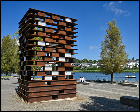 Rostfarbene Metallskulptur in Form eines durchbrochenen Turms am Seeufer. Die würfelartige Konstruktion mit horizontalen Ebenen erlaubt Durchblicke zur Wasserfläche und zur Wohnsiedlung im Hintergrund