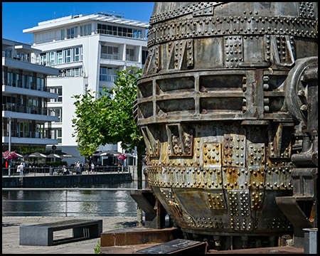 Nahaufnahme des historischen Stahlkonverters mit seinen charakteristischen Nieten und Metallbändern. Im Hintergrund sind moderne Wohngebäude und eine kleine Grünfläche zu sehen