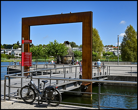 Großer rostfarbener Metallrahmen als Teil einer Brücke, der den Blick auf den See und die Wohnsiedlung im Hintergrund einrahmt. Im Vordergrund ein abgestelltes Fahrrad und eine Informationstafel.