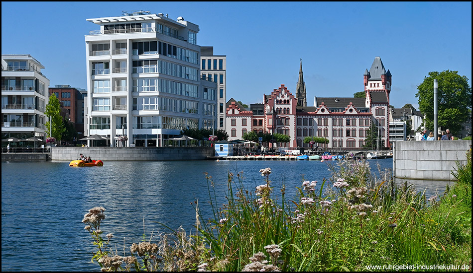 Uferpromenade mit üppiger Wildblumenwiese im Vordergrund. Über den See hinweg sieht man moderne weiße Wohngebäude und das historische rot-weiße Gebäude mit charakteristischem Turm. Ein gelbes Tretboot bewegt sich auf dem Wasser.