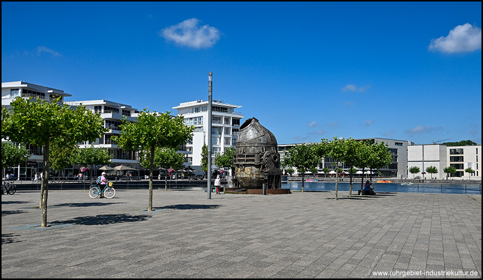Weiter Platz am Seeufer mit Metallskulptur des historischen Konverters in der Mitte. Die Fläche ist von jungen Bäumen umgeben, im Hintergrund stehen moderne weiße Wohngebäude. Eine Person fährt mit dem Fahrrad durch den Platz