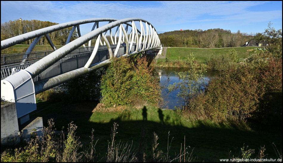 Weiße Stahlbogen-Brücke über einen Fluss im Herbst. Die Büsche sind bunt. 