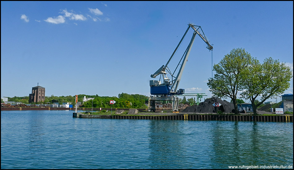 Ein großer blauer Hafenkran steht am Ufer eines Industriehafens. Im Hintergrund ist ein historischer Backsteinturm zu sehen. Das Wasser spiegelt den blauen Himmel, und am Ufer sind Bäume und Industriestrukturen erkennbar.