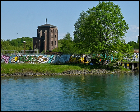 Ein alter Industrieturm aus rotem Backstein erhebt sich am Ufer des Kanals. Eine mit Graffiti bedeckte Mauer grenzt ans Wasser. Der klare blaue Himmel und das grüne Laub der Bäume bilden einen schönen Kontrast zur Industriekulisse.