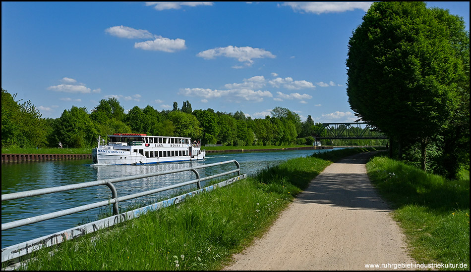 Ein weißes Ausflugsschiff fährt auf dem Kanal. Zu beiden Seiten des Wasserwegs erstrecken sich grüne Ufer mit Bäumen. Am Horizont ist eine Eisenbahnbrücke zu sehen, und rechts verläuft ein Wanderweg.