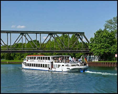 Ein Passagierschiff mit Touristen an Bord fährt unter einer Eisenbahnbrücke hindurch. Das Wasser glänzt in der Sonne, und im Hintergrund ist üppiges Grün zu sehen.