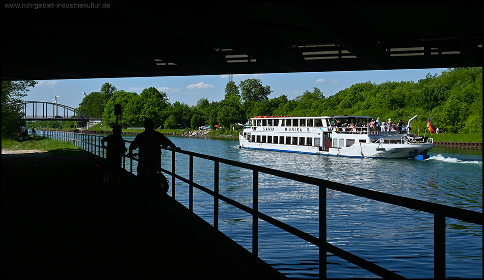 Zwei Silhouetten von Menschen beobachten vom Ufer aus ein Ausflugsschiff namens "Santa Monika II", das auf dem Kanal vorbeifährt. Die Szene wird durch den Rahmen einer Brücke gerahmt.