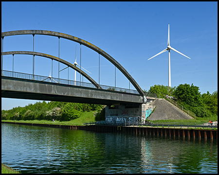 Eine Bogenbrücke überspannt den Kanal, dahinter ist ein großes Windrad zu sehen. Das klare Wasser reflektiert den strahlend blauen Himmel und die umliegende Landschaft.