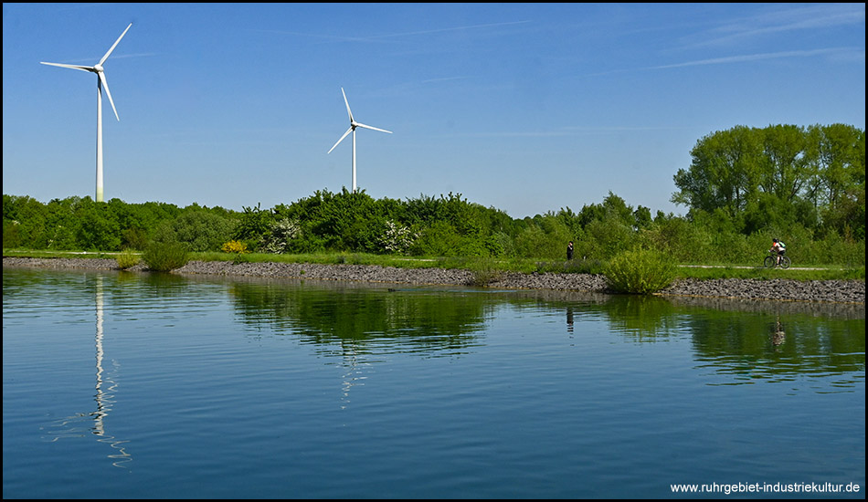 Zwei Windräder ragen über einer grünen Landschaft am Kanal auf. Ein einsamer Radfahrer ist am Uferweg zu sehen. Das ruhige Wasser spiegelt den blauen Himmel und die Umgebung.