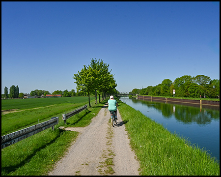 Eine Person in türkisfarbenem Oberteil radelt auf einem Uferweg neben einem Kanal. Links erstrecken sich grüne Wiesen und rechts das ruhige Wasser unter strahlend blauem Himmel. Eine Reihe junger Bäume säumt den Weg.