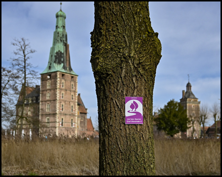 Wegmarkierung "Landstreifer" an einem Baum. Dahinter unscharf Schloss Raesfeld