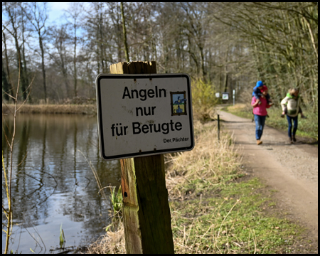Schild "Angeln nur für Befugte" an einem Seeufer, daneben Weg mit Wanderern 