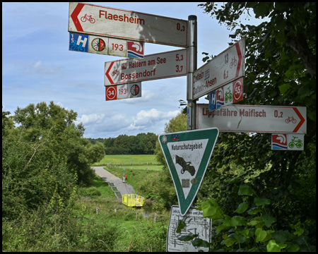 Wegweiser für Radfahrer. Im Hintergrund kleine Fußgängerfähre an einem Fluss