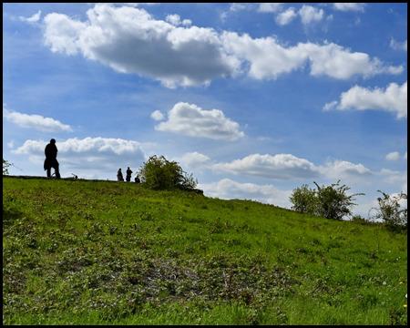 Eine grasbewachsene Böschung. Am Oberen Ende stehen Menschen als dunkle Figuren im Gegenlicht 