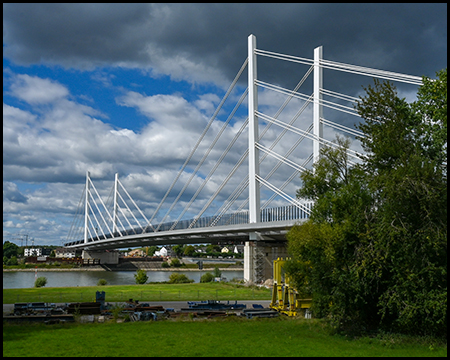 Moderne Schrägseilbrücke mit weißen Pylonen und Stahlseilen überspannt einen breiten Fluss, im Vordergrund grüne Wiese und Bäume unter dramatischem Wolkenhimmel.
