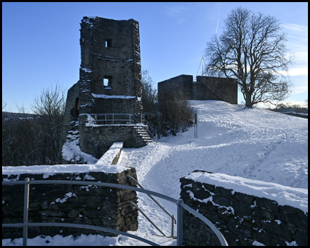 Gelände einer Burgruine im Schne mit einem Turm, mehreren Mauern und einem laublosen Baum in einer Fläche. 