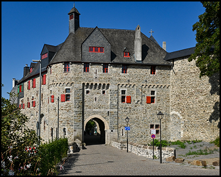 Eine Burgmauer aus Bruchstein mit Torbogen, roten Fensterläden und spitzen Dachgauben unter blauem Himmel.