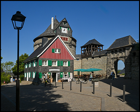 Ein Fachwerkhaus mit rot-weißer Fassade und Turmuhr neben einem gepflasterten Burgplatz mit Torbogen.