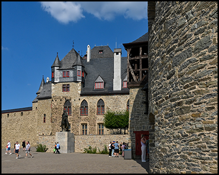 Ein Burghof mit Reiterstandbild vor einer Schlossfassade mit Türmchen, Besucher gehen über den Platz.