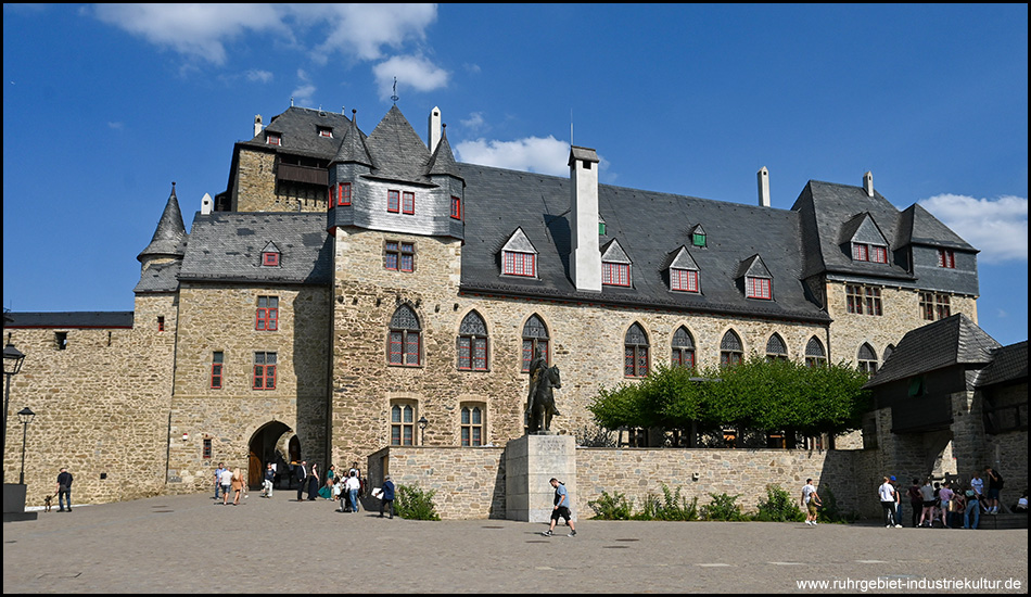 Die Schlossanlage mit Fachwerkgiebeln, Erkertürmen und Schieferdach, im Vordergrund ein Reiterdenkmal und Besucher auf dem Platz.
